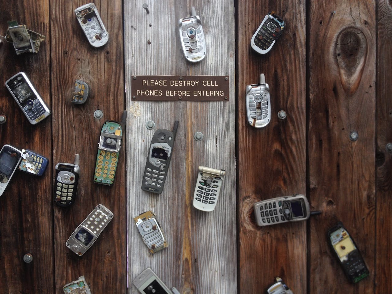 Collection of vintage cell phones attached to a rustic wooden wall with a humorous sign.